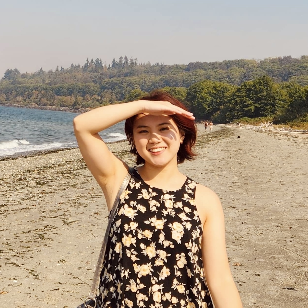 A casual photo of Sam from the waist up. She has short, reddish hair and a big smile. She's blocking the sun from her eyes and wearing a black dress with yellow and white flowers. She's in Discovery Park, right by the sound, and in the distance, there is lush greenery.